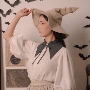 May include: A woman wearing a beige witch hat and a white blouse with a dark grey collar. The blouse has long, puffy sleeves. The background features black bat cutouts and the text "Stitch Maiden".