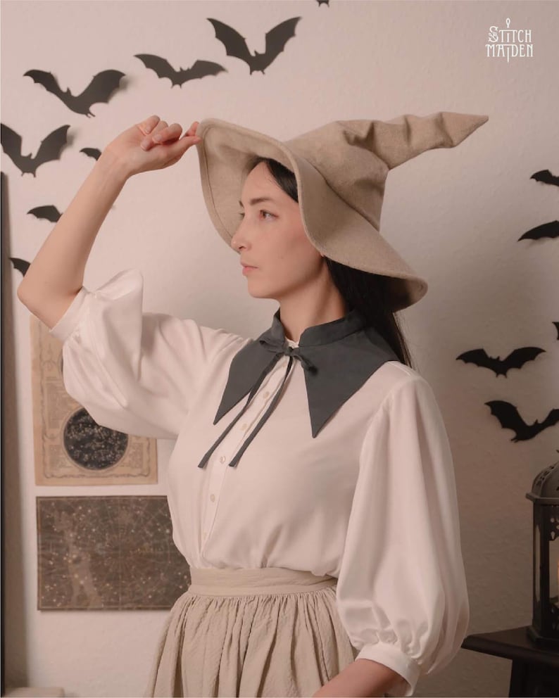May include: A woman wearing a beige witch hat and a white blouse with a dark grey collar. The blouse has long, puffy sleeves. The background features black bat cutouts and the text "Stitch Maiden".