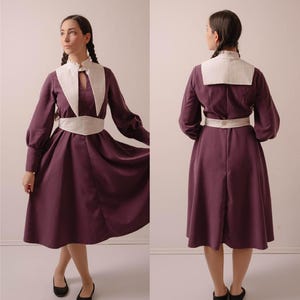 May include: A plum-coloured dress with long sleeves, a white collar, and a matching white belt. The dress has a keyhole neckline and a flared skirt. The sleeves are gathered at the cuffs. The model is wearing black flats. The dress is from Stitch Maiden.