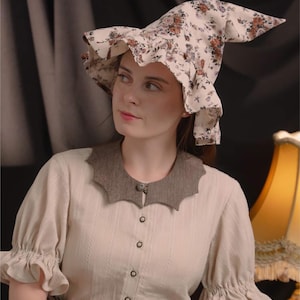 May include: A cream blouse with a brown, scalloped collar and button-down front. The blouse has short, ruffled sleeves. A floral print witch hat with a pointed crown and ruffled brim is worn.