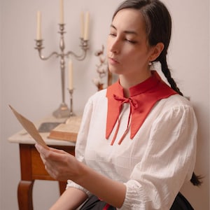 May include: A white blouse with a red collar and a black skirt. The red collar is tied with a bow. The blouse has long sleeves. The image also shows a silver candelabra and the text "STITCH MAIDEN".