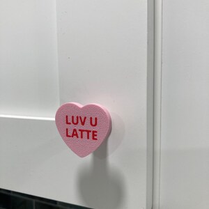 May include: A pink heart-shaped cabinet knob with the words "LUV U LATTE" in red lettering. The knob is mounted on a white cabinet door. The image is a close-up of the knob and part of the cabinet.