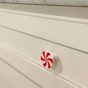 May include: A white cabinet drawer with a red and white peppermint swirl knob. The knob is round with a white base and a red spiral design. The cabinet is painted white with horizontal grooves.
