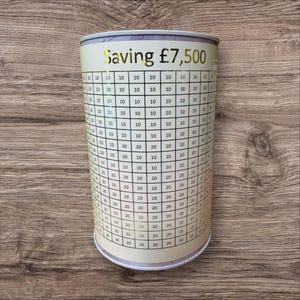 Könnte beinhalten: Eine weiße zylindrische Sparbüchse mit einem aufgedruckten Diagramm auf der Seite. Das Diagramm zeigt Zahlen von 10 bis 20 und den Text "Saving £7,500".