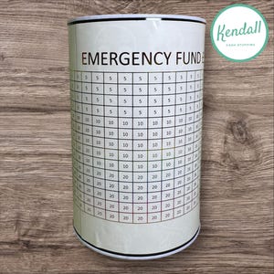 May include: A white cylindrical money jar with a chart on the side for saving money. The chart is labelled "Emergency Fund" and has numbers from 5 to 20. The jar has a black and white logo that says "Kendall Cash Stuffing".