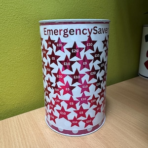 May include: A white cylindrical tin with a pink and white polka dot pattern around the bottom. The tin has a pink star pattern with the text "Emergency Saver" and various amounts of money in pounds sterling printed on the stars.