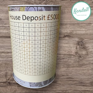 May include: A white cylindrical money box with a gold label that reads "House Deposit £5000". The box has a grid pattern with numbers from 5 to 20.
