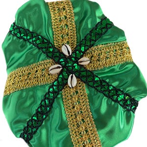 Op de afbeelding: Een groene satijnen bonnet met gouden en zwarte decoratieve randen. De bonnet heeft een kruisontwerp met gouden randen en zwarte pailletaccenten. Vier schelpen zijn in het midden van het kruis geplaatst.