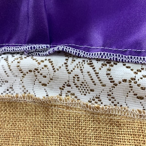 May include: Close-up of a purple fabric with white stitching, layered over white lace and brown hessian fabric.