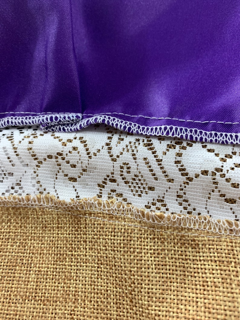 May include: Close-up of a purple fabric with white stitching, layered over white lace and brown hessian fabric.