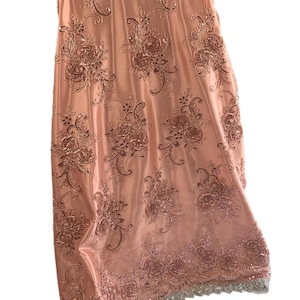 Puede incluir: Tela de color melocotón con un patrón floral y lentejuelas. La tela tiene un borde festoneado con un ribete plateado.