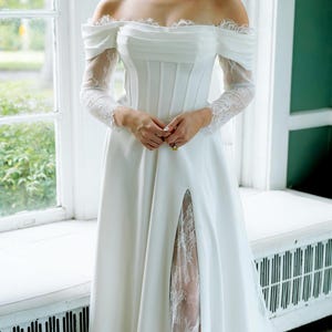 May include: Off-the-shoulder white wedding dress with long lace sleeves and a high slit revealing a lace panel. The bodice is fitted, and the skirt flows to the floor. The dress is worn in front of a window.