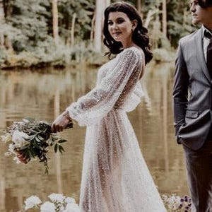 以下が含まれることがあります： 湖の前で結婚式の写真を撮るカップル。花嫁は長袖の白いビーズのドレスを着ており、新郎は暗いネクタイをつけたグレーのスーツを着ています。カップルは花と緑に囲まれた木製の桟橋に立っています。