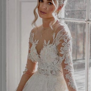 May include: A woman wearing a white lace wedding dress with a plunging neckline and long sleeves. The dress has a full skirt and is adorned with delicate floral embroidery. The woman is looking at the camera with a soft smile.