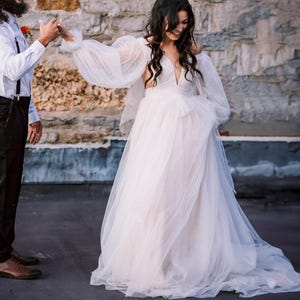 Pode incluir: Um casal posa para uma foto de casamento. A noiva está usando um vestido branco de tule com decote profundo e mangas grandes e bufantes. O noivo está usando uma camisa branca de botões, calças pretas e suspensórios.