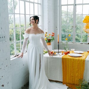 May include: A white off-the-shoulder wedding dress with long lace sleeves and a high slit. The dress is worn in a bright room with large windows and a table set for a meal with a yellow runner.