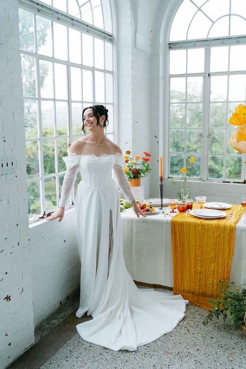 May include: A white off-the-shoulder wedding dress with long lace sleeves and a high slit. The dress is worn in a bright room with large windows and a table set for a meal with a yellow runner.