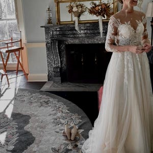 May include: A bride wearing a white lace wedding dress with a long tulle skirt and a floral design. She is standing in front of a fireplace with a mirror above it.