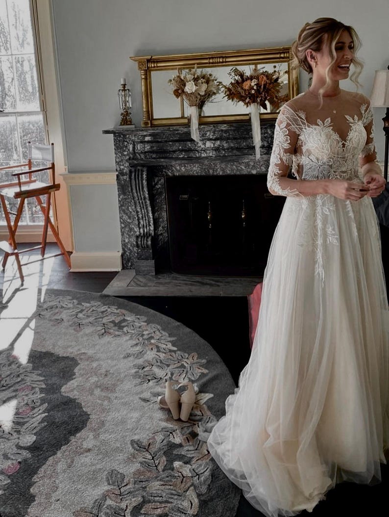 May include: A bride wearing a white lace wedding dress with a long tulle skirt and a floral design. She is standing in front of a fireplace with a mirror above it.