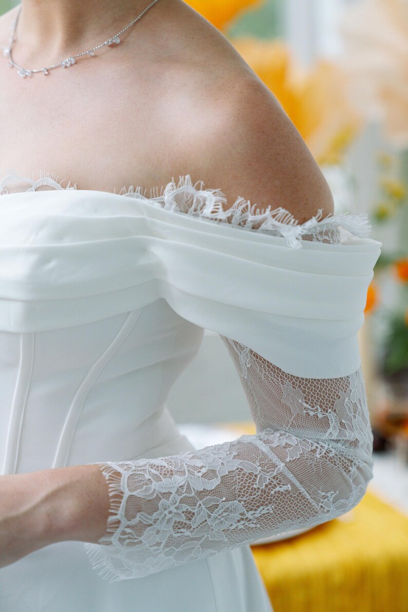 May include: A white off-the-shoulder wedding dress with lace sleeves and a delicate necklace. The dress features a draped bodice and lace trim along the neckline. The sleeves are sheer lace, adding a touch of elegance.