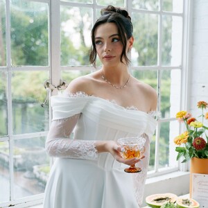 May include: A white off-the-shoulder wedding dress with lace sleeves and a high slit. The bride holds a glass of amber liquid. A table displays fruit and flowers, with a window in the background.