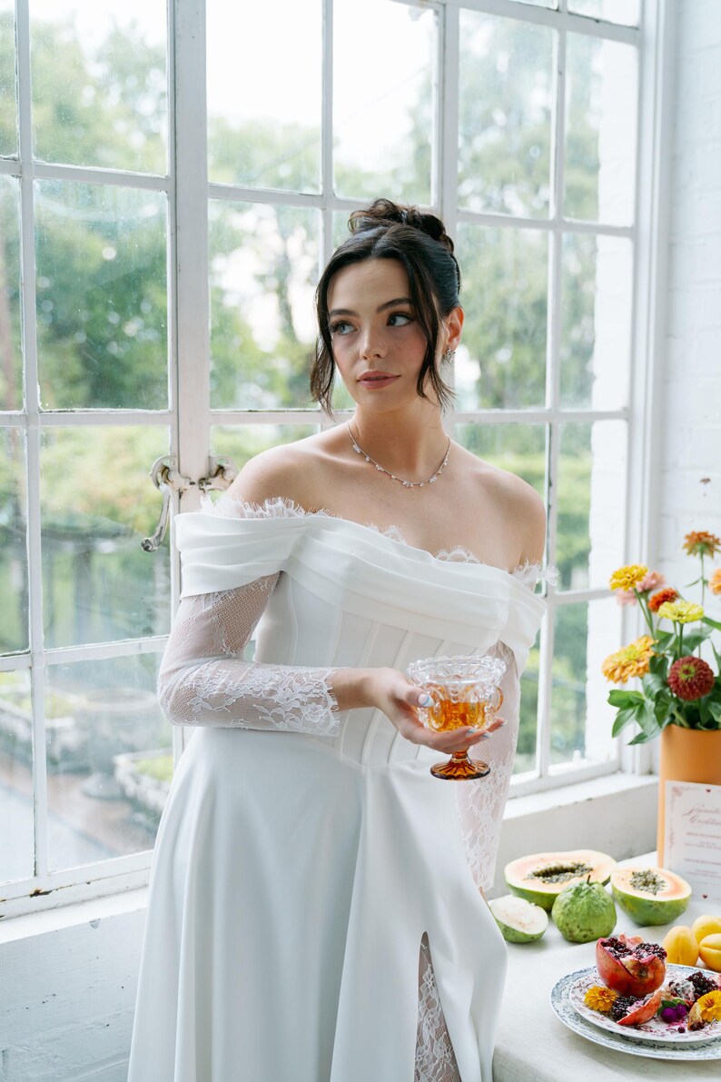 May include: A white off-the-shoulder wedding dress with lace sleeves and a high slit. The bride holds a glass of amber liquid. A table displays fruit and flowers, with a window in the background.