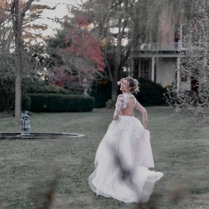 May include: A bride in a white lace wedding dress with a low back and long sleeves walks away from the camera in a garden setting. The dress has a flowing skirt and a train. The bride is smiling and looking over her shoulder.