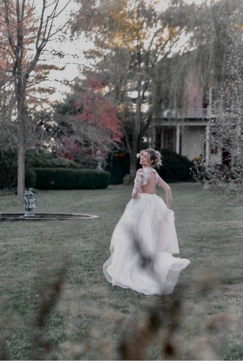 May include: A bride in a white lace wedding dress with a low back and long sleeves walks away from the camera in a garden setting. The dress has a flowing skirt and a train. The bride is smiling and looking over her shoulder.