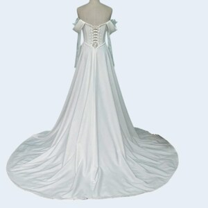 May include: A white wedding dress with an off-the-shoulder neckline and a long train. The back features a corset-style closure. The sleeves are long and flowing, and the dress is displayed on a mannequin.