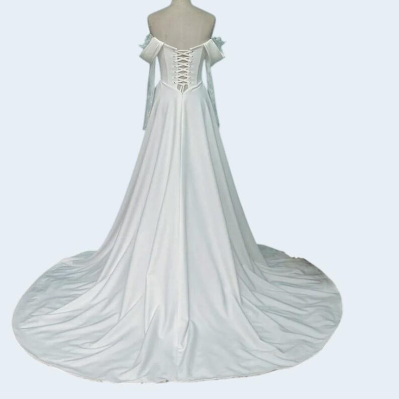 May include: A white wedding dress with an off-the-shoulder neckline and a long train. The back features a corset-style closure. The sleeves are long and flowing, and the dress is displayed on a mannequin.