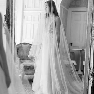 May include: A bride wearing a white wedding dress with a long veil. The bride is standing in front of a mirror and looking at her reflection.