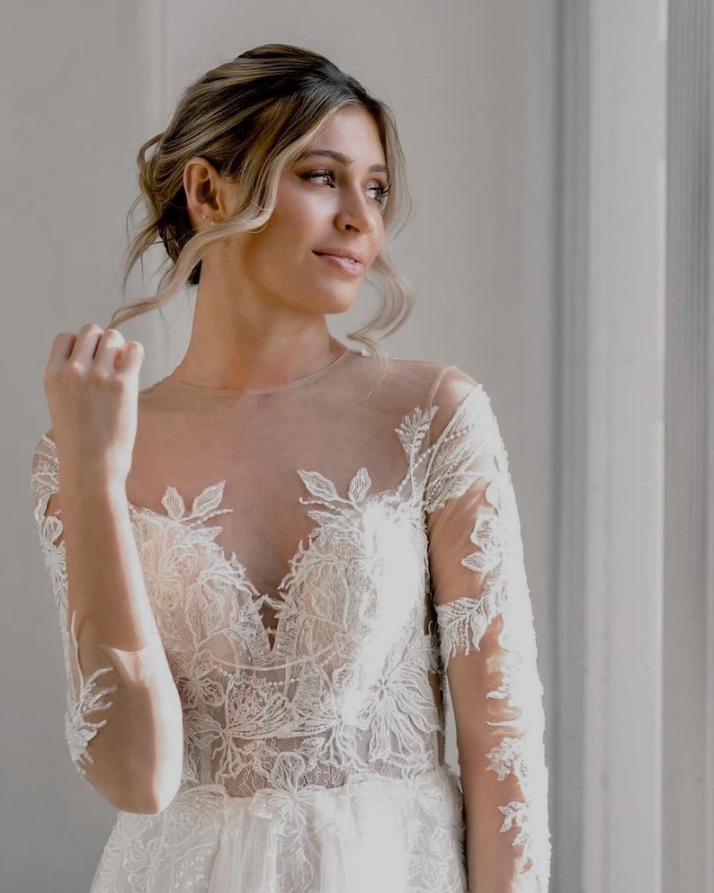 May include: A bride wearing a white lace wedding dress with a sheer bodice and long sleeves. The dress has a fitted bodice and a flowing skirt. The bride is looking off to the side with a soft smile.
