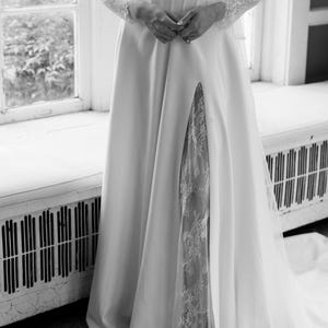 May include: A white wedding dress with a high slit revealing a lace panel. The dress features a fitted bodice, off-the-shoulder sleeves, and a flowing skirt. The long sleeves are made of lace. The dress is elegant and suitable for a wedding.