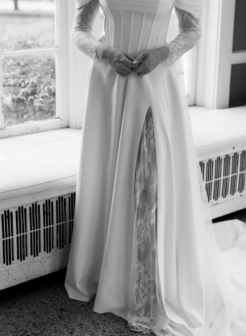 May include: A white wedding dress with a high slit revealing a lace panel. The dress features a fitted bodice, off-the-shoulder sleeves, and a flowing skirt. The long sleeves are made of lace. The dress is elegant and suitable for a wedding.