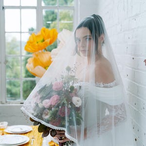 May include: A bride wearing a white off-the-shoulder wedding dress and a sheer veil. She holds a bouquet of pink and white flowers with greenery. The veil is trimmed with small white beads. Large yellow paper flowers are in the background.
