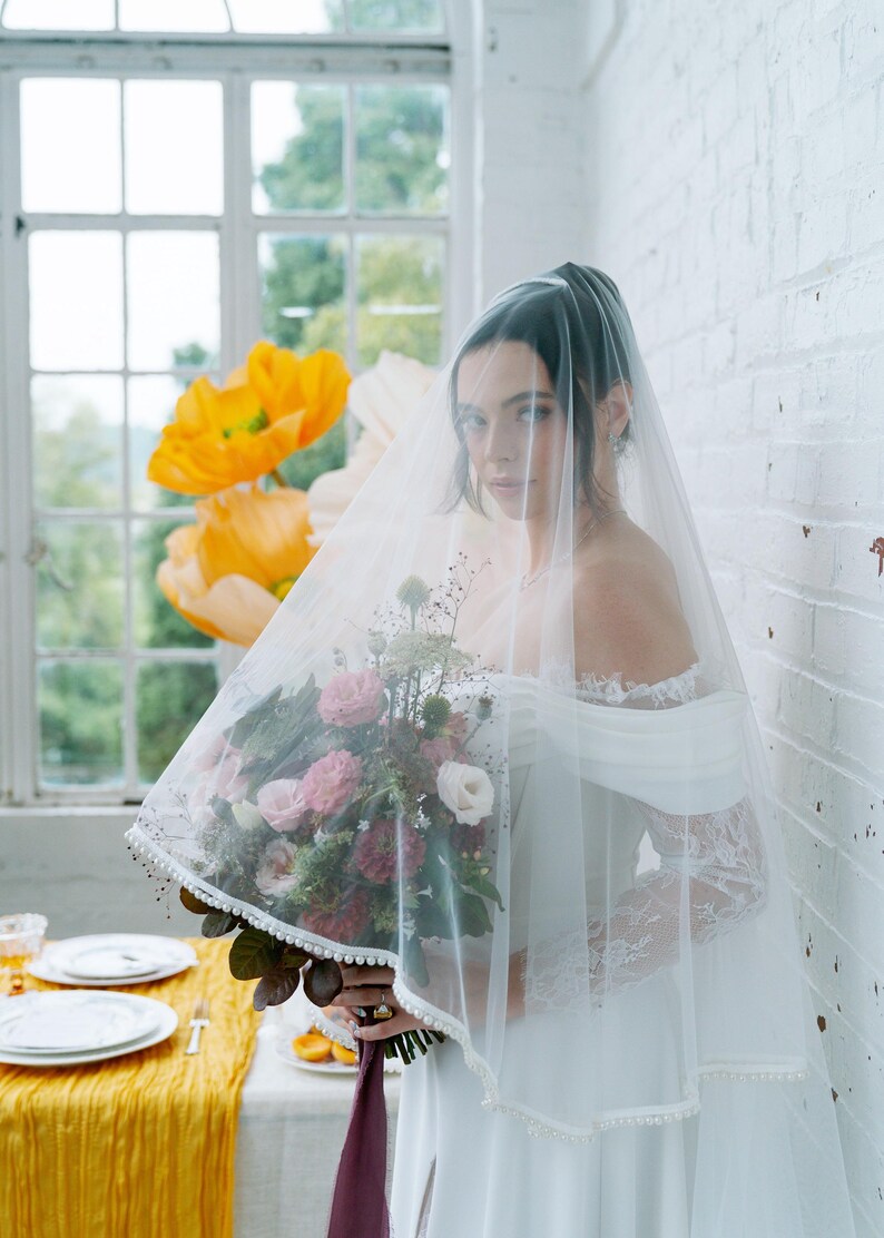 May include: A bride wearing a white off-the-shoulder wedding dress and a sheer veil. She holds a bouquet of pink and white flowers with greenery. The veil is trimmed with small white beads. Large yellow paper flowers are in the background.