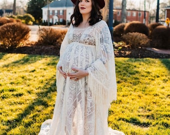 May include: A pregnant person wearing a white lace maxi dress with fringe details and a burgundy wide-brimmed hat. The dress is sheer and has a flowing skirt. The person is standing in a grassy field.