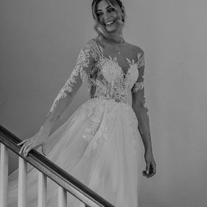 May include: A bride in a white lace wedding dress with a tulle skirt, smiling as she walks down a staircase.