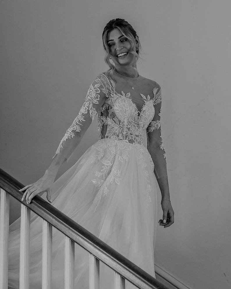 May include: A bride in a white lace wedding dress with a tulle skirt, smiling as she walks down a staircase.
