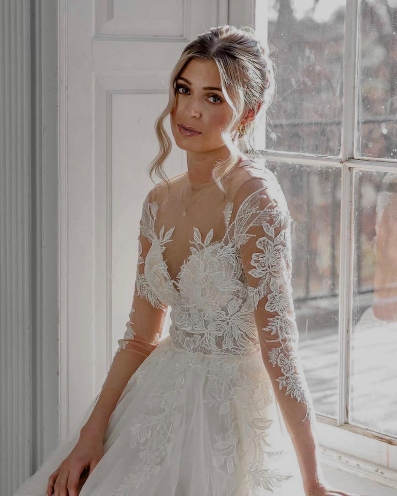 May include: A woman wearing a white lace wedding dress with a plunging neckline and long sleeves. The dress has a full skirt and is adorned with delicate floral embroidery. The woman is looking at the camera with a soft smile.