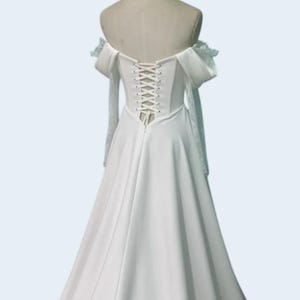 May include: A white off-the-shoulder wedding dress with a corset back. The dress features long, sheer sleeves with lace detailing and a flowing skirt. The bodice has a lace-up closure, and the dress is displayed on a mannequin.