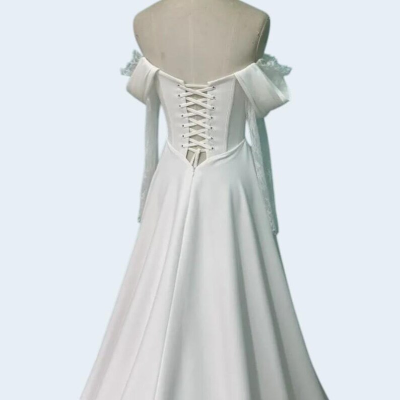 May include: A white off-the-shoulder wedding dress with a corset back. The dress features long, sheer sleeves with lace detailing and a flowing skirt. The bodice has a lace-up closure, and the dress is displayed on a mannequin.