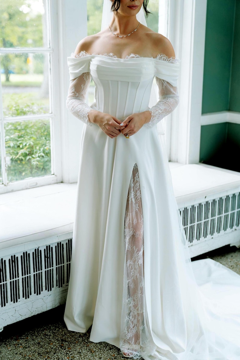 May include: Off-the-shoulder white wedding dress with long lace sleeves and a high slit revealing a lace panel. The bodice is fitted, and the skirt flows to the floor. The dress is worn in front of a window.
