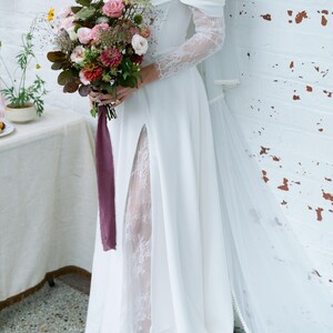 May include: A white off-the-shoulder wedding dress with lace sleeves and a high slit. The bride holds a bouquet of pink, red, and green flowers tied with a purple ribbon. A sheer veil drapes down the back.