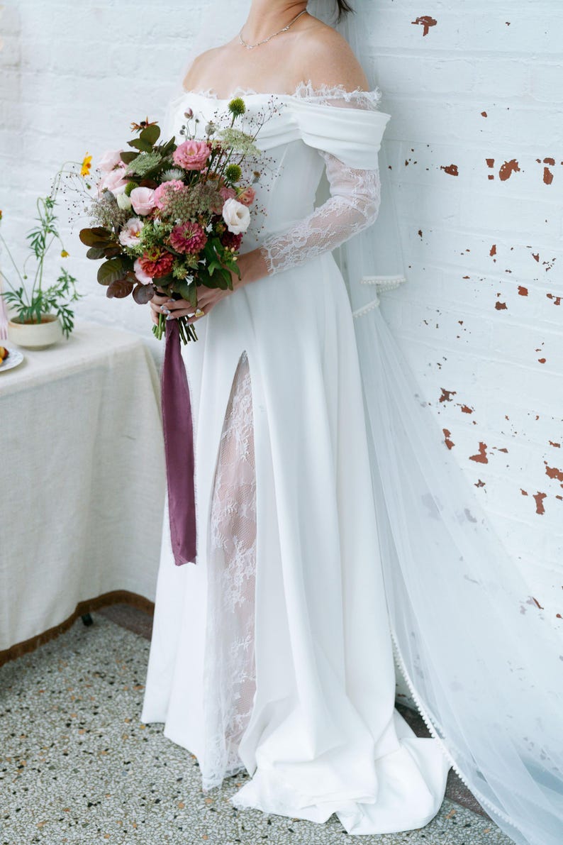 May include: A white off-the-shoulder wedding dress with lace sleeves and a high slit. The bride holds a bouquet of pink, red, and green flowers tied with a purple ribbon. A sheer veil drapes down the back.