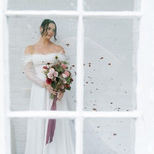 May include: A bride in an off-the-shoulder white wedding dress with lace sleeves holds a bouquet of pink, red, and green flowers tied with a burgundy ribbon. The bride stands in front of a white brick wall, seen through a window.
