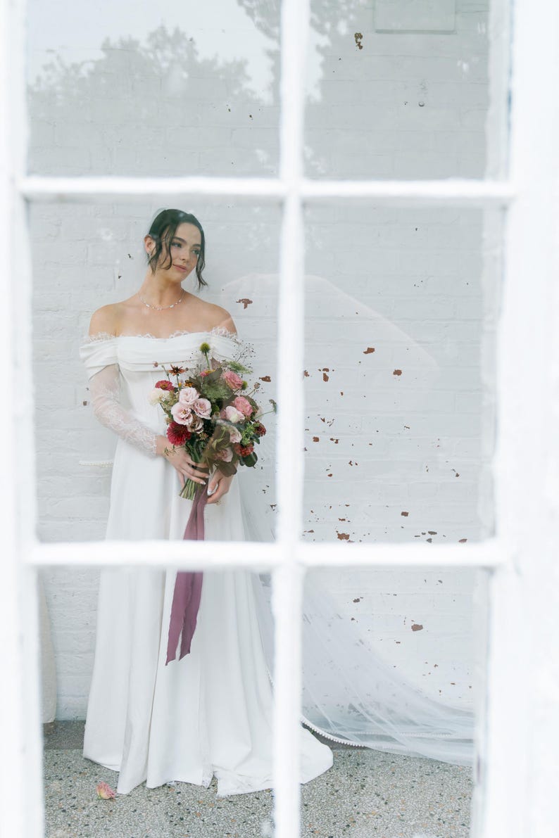 May include: A bride in an off-the-shoulder white wedding dress with lace sleeves holds a bouquet of pink, red, and green flowers tied with a burgundy ribbon. The bride stands in front of a white brick wall, seen through a window.