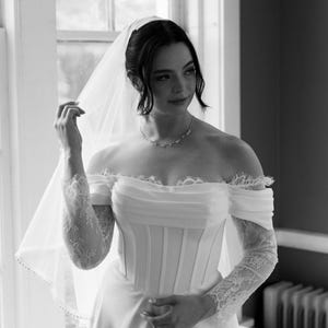 May include: A bride in a white off-the-shoulder wedding dress with lace sleeves and a veil. The dress features a fitted bodice and a flowing skirt. The bride is wearing a delicate necklace.