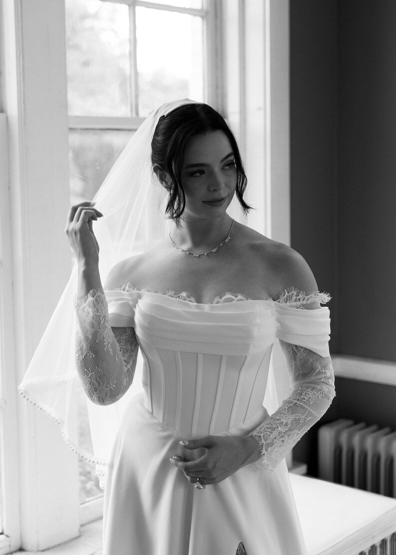 May include: A bride in a white off-the-shoulder wedding dress with lace sleeves and a veil. The dress features a fitted bodice and a flowing skirt. The bride is wearing a delicate necklace.