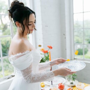 May include: A white off-the-shoulder dress with lace sleeves. A woman is holding a white plate with a blue floral design. The table is set with plates, glasses, fruit, and flowers. The scene is set in front of a large window.
